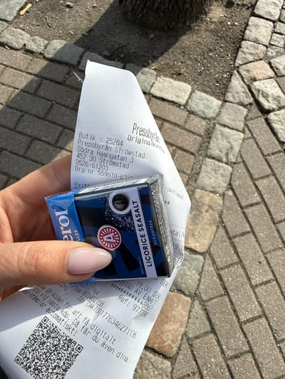 An overhead outdoor shot captures a hand holding a receipt and a small box of candy against a background of grey cobblestones and paved ground. The scene is brightly lit by natural sunlight, indicating daytime.
The receipt is from "Pressbyrån" and is labeled "Originalkvitto." Visible text details include "Butik : 25284 Pressbyrån Strömstad Södra Hamngatan 3 452 30 Strömstad Org nr 559010-632." The receipt shows a transaction of "19,00 SEK" and mentions a "Ref: 977998." It also features a QR code and instructions in Swedish: "Spara denna QR-koden för att få digitalt kvitto vid nästa köp. Då får du även dina app-stämplar automatiskt!" (Save this QR code to get a digital receipt for your next purchase. Then you will also get your app stamps automatically!).
The small, blue and black box of candy is branded "erol" and clearly displays "LICORICE SEASALT" and "SUGARFREE." It also shows a net weight of "25 g" and mentions "PL 406, 20101 Turku, Finland," indicating manufacturing or distribution information. A circular logo with a letter "A" is prominent on the box. The hand holding the items has light pink manicured nails. The setting is likely a street or public area in Strömstad, Sweden, as indicated by the receipt details.