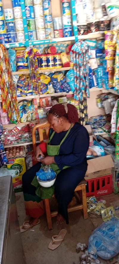 This image captures the interior of a small, well-stocked shop, likely a local convenience store, in Gayawa, Nigeria. The scene is dominated by a woman, appearing to be middle-aged, with red dreadlocks tied in a ponytail. She is wearing a dark blue long-sleeved top and a green apron, and is seated on a simple wooden stool.

The woman is actively engaged, looking down at her hands, where she holds a small item over a white bowl that contains a blue plastic bag. This suggests she might be weighing or packaging goods for a customer. Her light-colored flip-flops are visible on the concrete floor beside her.

The shop itself is densely packed with various products arranged on wooden shelves. The top shelves are filled with numerous tins of milk powder, identifiable by brands such as "NAN" and "PUREMILK." A shelf divider prominently displays the text "BREMFAST TIME." Lower shelves showcase an array of packaged items, including yellow bags of "Chelsea" sugar, "SUPREME NOODLES" with "TASTY CHICKEN" flavor, and other household goods. Bundles of sachets hang from strings, adding to the cluttered yet functional appearance.

On the left side of the frame, a tray of eggs is visible, along with a package labeled "CHERIE 20g." The floor on the right features a red "Coca-Cola" crate and a large blue plastic bag with the brand name "JEANIE," surrounded by some scattered wrappers. The overall impression is that of a bustling, essential local business. The lighting is ambient, with no distinct time of day or weather indicated.