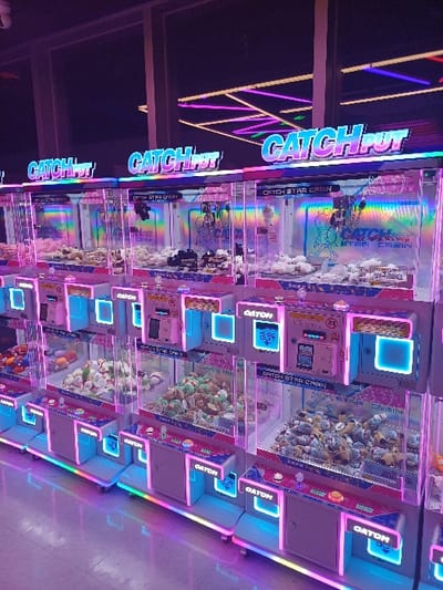 The image depicts an interior view of an arcade in 마포구, South Korea, focused on a dense arrangement of claw game machines. There are no individuals present. Multiple "CATCH PUT" and "CATCH STAR CABIN" units, illuminated by neon pink, purple, and blue LED lighting, are arranged in rows. Each machine features a transparent front showcasing a variety of plush toys, including white, green, brown, and assorted multi-colored characters. The control panels on the lower section of each machine are brightly lit with blue and pink accents. The bases of the machines are equipped with multi-colored LED strips that reflect onto a light-colored tiled floor. The background is darker, with linear neon light fixtures visible on the ceiling, suggesting an enclosed commercial space.