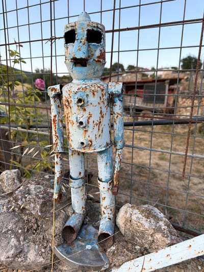 A weathered, pale blue and rusty metal robot sculpture stands in front of a wire fence in Salmerón, Spain. The robot has a cylindrical body with three visible bolt-like circles on its chest and articulated arms and legs made from pipes. Its head is blocky with large, dark rectangular eye sockets and a small, rectangular mouth. The robot is positioned on a metal circular base that rests on two large rocks. Behind the robot, the wire fence is visible, with vegetation and buildings in the background under a clear sky. A pink rose is blooming on a vine to the left of the robot, partially obscured by the fence. The environment appears to be outdoors, possibly in a rural or semi-rural setting within the town.