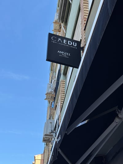 The image depicts an upward angle view of a building facade in L'Alcúdia de Crespins, Spain, under a dark awning. The sky is clear blue. A black rectangular sign is attached to the awning. The sign displays "CAEDU lapos-enirans-brasa" and the "AMSTEL" logo with "Hecha en Valencia" written below. Air conditioning units are affixed to the building exterior. The building's architectural details include light-colored brickwork and varying paint colors. Structural metal supports hold up the awning.