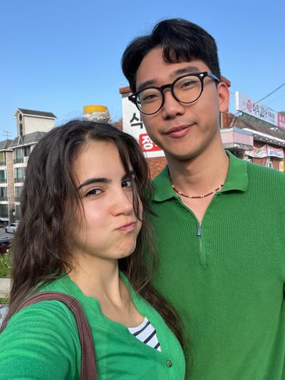 The media file is a close-up, selfie-style portrait of two individuals, a man and a woman, against a backdrop of buildings and a clear blue sky. The image is oriented sideways, as if taken from a phone held vertically.
The man is on the left, wearing black-rimmed glasses and a green collared shirt with a zipper detail. He has dark hair and is smiling gently. A beaded necklace with red and white beads is visible around his neck.
The woman is on the right, with long, dark, wavy hair. She is also wearing a green top, which has a ribbed texture and appears to be a cardigan over a striped shirt. Her expression is neutral, with her lips slightly parted.
The background shows buildings, some with signage that includes Korean characters. One sign clearly reads "RO" in large red letters. There are also hints of streetlights and overhead wires, suggesting an urban or semi-urban environment. The clear blue sky indicates it is daytime, likely during fair weather. The overall impression is a casual, candid moment captured between two people, possibly friends or a couple, in South Korea.
