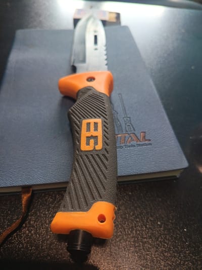 The image shows a close-up of an orange and black handled knife resting on a blue notebook. The notebook has a textured cover and appears to have some text and a graphic printed on it. The knife has a serrated blade and a black handle with a textured grip. The overall scene is lit from above, casting shadows. The location in Kapan, Armenia, cannot be determined from the provided image.