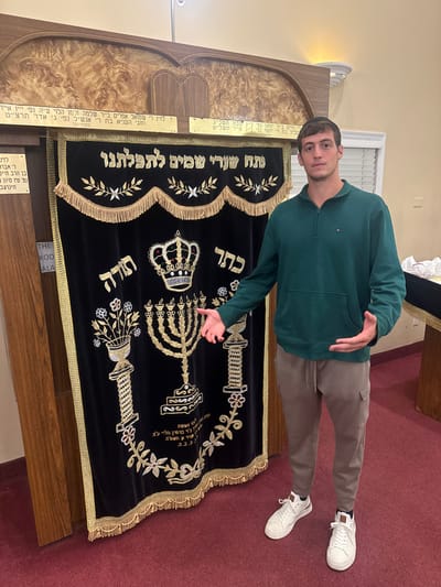 The image displays a young adult male inside a synagogue in Bergenfield, United States. The subject stands slightly right of center, facing the camera, with an elaborate Aron Kodesh (Holy Ark) directly behind him and to his left.
The male, appearing to be in his early twenties, has short, dark hair and is clean-shaven. He wears a dark teal quarter-zip pullover sweatshirt with a small brand logo on the left chest, light brown cargo-style jogger pants, and white sneakers with brown accents. His arms are bent, and his open hands gesture towards the Aron Kodesh.
The Aron Kodesh is constructed of light-toned wood with an arched top and features two plaques bearing Hebrew inscriptions above a dark velvet curtain (parochet). The curtain is heavily embroidered in gold and silver thread, depicting a large crown flanked by the Hebrew words "כתר" (Crown) and "תורה" (Torah), a prominent seven-branched menorah, and ornate vases with floral arrangements atop columns. Additional Hebrew text is embroidered across the top section of the curtain, and gold fringe borders its top and bottom edges.
The interior environment features light-colored walls and a red carpeted floor. A wall sconce is visible on the upper right wall, and a window with white blinds is present in the background to the right. A partially visible wooden structure covered with a white cloth and some white bags are also present on the far right.