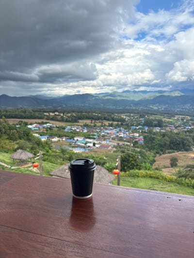 A black ribbed paper coffee cup with a black plastic lid and white exposed rim is centered on a dark brown wooden surface in the foreground. The image presents an elevated panoramic view of the area identified as the city of Ban Mai Saha Sampan, Thailand. Below the wooden surface, a steep, verdant hillside slopes downwards, revealing a settlement comprising numerous structures with multi-colored roofs (predominantly blue, red, grey) situated amidst trees. Two circular, thatched-roof huts and two red spherical lanterns are visible on this slope. Beyond the immediate settlement, a wide valley extends towards a distant, undulating mountain range covered in dense vegetation. The sky overhead is heavily overcast with large, dark grey cloud formations, though patches of lighter clouds and blue sky are discernible towards the upper right. No persons are depicted within the scene.