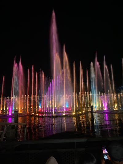 An illuminated fountain display is visible at night in what is identified as Pardomuan I, Indonesia. Multiple water jets, lit with vibrant pink, purple, orange, and blue lights, arch upwards from a body of water. The colored lights reflect on the dark surface of the water. In the foreground, the tops of two people's heads are visible, suggesting they are watching the display. One person is holding a smartphone, possibly to take a picture or video. A dark railing separates the viewers from the fountain area. The background is a solid black sky.