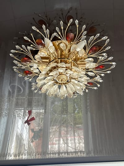 An interior scene in Dmitriyevskaya, Russia, displays a prominent, ornate ceiling light fixture. The flush-mounted fixture features a gold-colored metallic frame, designed with radiating arms that hold numerous clear, faceted crystal pendants, some teardrop-shaped and others pointed. Interspersed among these are larger, deep red teardrop-shaped crystal pieces. At least one frosted white bulb is visible within the fixture, which is affixed to a light-colored, glossy ceiling, reflecting the fixture prominently. Below and to the left of the light fixture, a window is covered by sheer, light-colored lace curtains with embroidered patterns along the bottom edges. Through the translucent curtains, an outdoor environment with trees or shrubs and indistinct background structures is visible. Affixed to the window pane itself are two decorative items: a red, irregular shape resembling a stylized "K" or an arrow, and a partially obscured green, stylized winged creature. No individuals or specific actions are depicted.