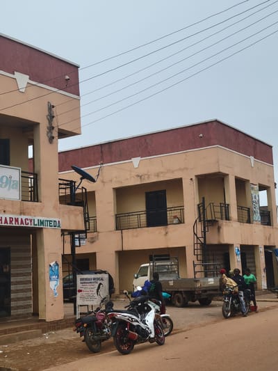 The image depicts a street scene in Tangu, Nigeria, with a multi-story building dominating the frame. The building is a faded peach color with maroon accents on the upper floors and along the roofline. Several balconies with black metal railings are visible on the building's facade, some containing objects. A satellite dish is mounted on one of the balconies. Power lines crisscross the sky above.

In the foreground, several motorcycles are parked along the side of the building, some with riders seated or standing nearby. A flatbed truck is also visible. People are interacting around the motorcycles and the truck, suggesting a moment of waiting or preparation for travel. One person is wearing a bright green shirt, and another has a patterned cloth wrapped around their head.

Several signs are posted on the building. One vertical sign reads "PHARMACYLIMITED". Another sign, with a silhouette of a woman, displays text including "BWEHMASI MEDICAL RADIDIAGNOSTIC SERVICES LTD" and a motto, "IN DAND WE TRUST". A smaller poster with a face and the word "LUKA" is also visible. The ground is a reddish-brown, characteristic of a dirt or unpaved road. The sky is overcast, suggesting a cloudy or possibly humid day. The overall impression is one of everyday life and commerce in a Nigerian town.