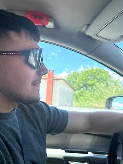 A man with short black hair and a goatee is seen from the side, driving a car. He is wearing black Prada sunglasses with a red stripe and a dark grey t-shirt. The interior of the car is visible, including the sun visor and a small red object with white text attached to the headliner. The view outside the car window shows a bright blue sky with some clouds, a house with an orange chimney, and lush green trees. A side-view mirror reflects the sky. The image appears to have been taken on a sunny day in Kato Nevrokopi, Greece.