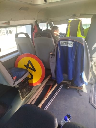 Inside a parked van in Manhush, Ukraine, a driver wearing glasses and a dark shirt is seated at the steering wheel, looking forward. A bright yellow safety vest is draped over the headrest of the passenger seat beside him. In the foreground, a circular road sign with a red border and a black number "4" on a yellow background is leaning against a seat. Adjacent to the sign are several tools, including an axe, a shovel, and what appear to be metal rods, laid out on the van's floor. A blue jacket with a white and black logo that reads "ЕЛТ ЗА" and some text below it is hanging from a makeshift stand in the middle of the van. A partially filled plastic bottle of water with a blue cap is visible on the floor near the back seats. The van's interior is utilitarian, with grey seats and a visible metal interior frame. The overall lighting suggests it is daytime.