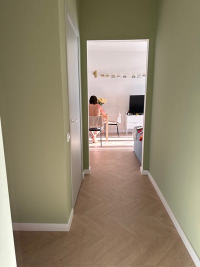 The image presents a view from an interior space, likely an entryway or hallway, looking into another room. The foreground features a doorframe, painted in a light green color. Beyond the doorway, the room has white walls and light wood-toned flooring with a chevron pattern. In the room, a person is seated at a small table or desk, and there are flowers on the table. A television is mounted on the wall in the background. The image is taken from an elevated perspective. It is not possible to determine the location in the city of Люк, Russia.