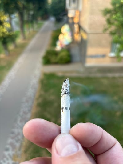 A close-up, shallow depth of field shot shows a person's thumb and index finger holding a partially smoked cigarette. The cigarette is white with a silver band and has ash on the tip, with wisps of smoke rising. The background is blurred, depicting a paved walkway lined with grass and trees, with a building visible in the distance. The scene appears to be outdoors in a residential area of Pereiaslav, Ukraine.