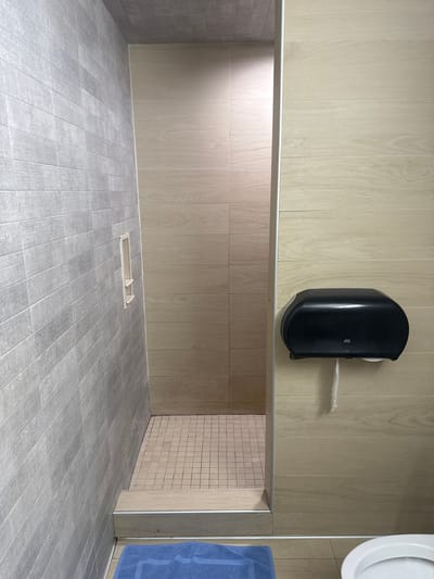 The image shows a small, tiled shower stall. The walls of the stall are tiled with light-colored, wood-grain patterned tiles, and the floor is tiled with small, square, beige tiles. The stall has a step leading up to it, and a blue mat is visible in front of the step. To the right of the shower stall, there is a black paper towel dispenser mounted on a wall tiled with the same light-colored, wood-grain tiles. A portion of a white toilet bowl is visible in the bottom right corner of the image. The overall impression is of a clean and functional, albeit basic, bathroom facility. The location is likely a public restroom or a gym changing room, possibly within the city of Burbank, United States, based on the context provided.