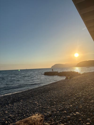 This image depicts a sunset scene on a pebble beach in Shirokaya Balka, Russia. The sun is setting over the sea, casting a warm golden glow on the water and the surrounding landscape. Several people are visible on the beach and on a pier, some sitting and observing the sunset, while others stand. A log lies on the beach in the foreground. The sky is clear blue with a hint of orange near the horizon. The sea is calm with gentle waves lapping the shore. Distant hills are visible on the horizon.