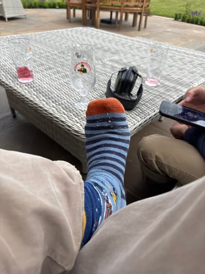 The image shows a person's foot wearing a blue and orange striped sock with a design on the bottom of the foot. The person is sitting on a patio in Sand Hutton, United Kingdom, which is located at latitude 54.025 and longitude -0.933. There is a wicker table in front of them with two glasses on it. The person is facing another person who is out of frame. The other person is holding a cell phone. The table is surrounded by patio furniture and a green lawn. The image is taken from a low angle looking up at the person's foot. The lighting is bright and sunny.