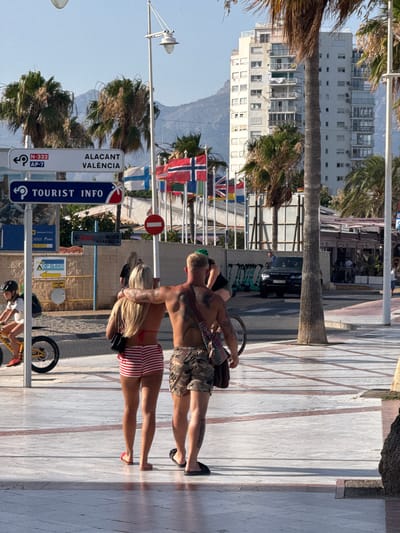 The image depicts a street scene in l'Alfàs del Pi, Spain, likely along a promenade. In the foreground, a man and a woman stand facing away from the camera. The man has tattoos on his back and arms and is wearing camouflage shorts. The woman has blonde hair and is wearing a red and white striped bikini top and shorts. They are standing on a paved walkway with patterned tiles.

Behind them, a young girl on a yellow bicycle rides past. Further in the background, there are traffic signs, including one pointing to "Alacant Valencia" and another indicating "Tourist Info". Various national flags are displayed on poles. Palm trees are visible, along with a tall, white apartment building in the distance. A black Range Rover is parked on the street, and several electric scooters are lined up. The scene is illuminated by sunlight, casting shadows on the ground.