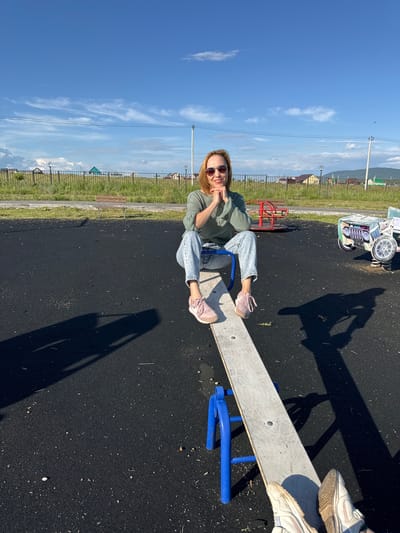 A woman is sitting on a seesaw on a playground in Mursalimkino, Russia. She is wearing a green hoodie, light blue jeans, and pink sneakers. She is smiling and looking at the camera, with her legs propped up on the seesaw. The seesaw is made of a grey metal plank and blue metal supports. The playground surface is black asphalt. In the background, there is a green grassy area with a fence and some houses. The sky is blue with some white clouds. The sun is shining, suggesting it is daytime. There is a shadow of a person on the seesaw, indicating the presence of another person or a camera operator. Other playground equipment is visible in the background, including a red structure and a white and green ride-on toy resembling a car. The scene conveys a sense of fun and relaxation.