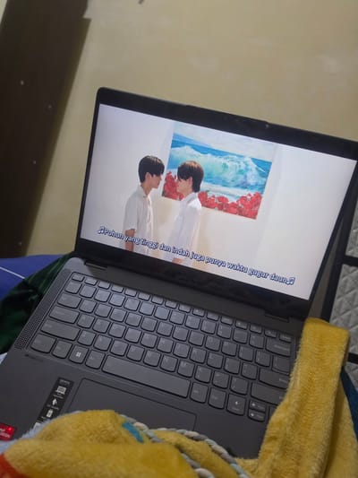 Here's a description of the image:
The image depicts a gray Lenovo laptop sitting on what appears to be a bed. The laptop screen is on and displaying a video, showing two men in front of an artwork depicting an ocean scene with red flowers. Subtitles in Indonesian are displayed on the screen. A yellow towel is partially draped over the keyboard of the laptop. The background is slightly out of focus but appears to be a wall. The image is taken indoors, with a slight diagonal orientation. I do not have information to ascertain the location of this image in Pintomakmur, Indonesia.