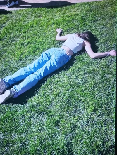 In Kasagaran, Uzbekistan, a single individual is depicted lying prone on a surface of green grass. The subject, with dark, wavy hair spread around the head, is dressed in a light-colored crop top or tank top, light blue denim jeans, and light-colored sneakers. The arms are extended laterally from the shoulders with palms facing downwards, and the legs are extended straight. A light-colored band is visible on the right wrist. In the upper left portion of the image, partial shadows of another person's legs and feet are visible, cast upon a darker surface adjacent to the grass. The scene is outdoors under bright sunlight.