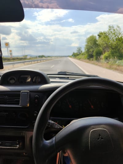 The image shows a view from inside a car driving on a highway. The steering wheel and dashboard are in the foreground, with the road stretching out ahead. The sky is blue with scattered clouds, and there are trees and fields on either side of the road. There are also signs for Shell gas stations visible in the distance. The overall impression is one of a pleasant drive on a sunny day.