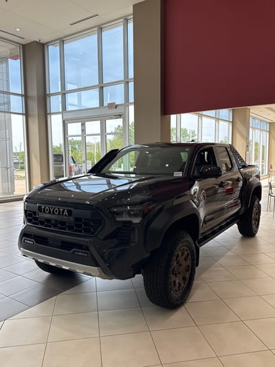 The image depicts a dark-colored Toyota truck, possibly a Tacoma TRD Pro, on display inside a showroom with large windows. The setting appears to be a car dealership with tiled flooring and interior architecture featuring red and beige walls. Natural light floods the scene through the windows. The location is likely in Palatine, United States of America. There are no people visible in the foreground of the image.