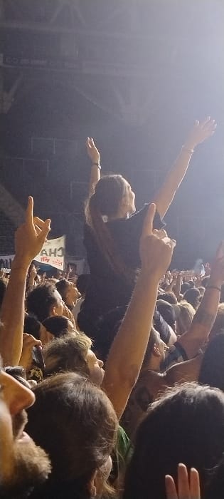 This image captures an energetic scene at a concert or live music event in Tolosa, Argentina. A dense crowd of people fills the foreground and mid-ground, with many individuals visible from the shoulders up, looking towards a presumed stage on the left. Numerous hands are raised in the air, some forming rock-and-roll gestures, conveying enthusiasm and engagement.
In the mid-ground, a person is seen crowd-surfing, lying on their back with arms outstretched, being carried above the audience's heads. This individual is wearing a dark t-shirt. Bright spotlights from the background create a hazy atmosphere and strong backlighting, highlighting the silhouetted figures and adding to the dramatic effect.
Partially visible behind the crowd-surfer, a vertical banner or sign displays text. The upper part features "CHIVA" in large white letters on a blue background, and below it, "ROCK Y BLUES" is written in red letters on a white background. The overall impression is one of a vibrant night-time musical performance, with the audience actively participating.
