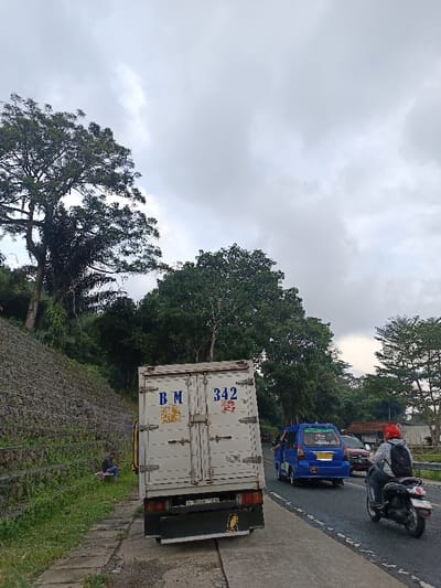 The image depicts a roadside scene in Cugenang, Indonesia, under an overcast sky. In the foreground, a white box truck is parked partially on a concrete sidewalk and partially on an asphalt road, angled towards the right. The truck's rear doors display "B M" on the left and "342" on the right in blue and red lettering, respectively. Its license plate, "D 9068 TH," is visible. To the immediate left of the truck, a vegetated embankment supported by gabion walls ascends steeply. At the base of this embankment, a person is seated on the grass beside the concrete path, facing the slope. On the asphalt road to the right of the truck, traffic is visible. A blue minivan is proceeding away from the viewer, followed by a dark-colored scooter with a rider wearing a red helmet and a dark backpack. Further down the road, a red vehicle is partially visible. The background consists of dense green foliage and numerous trees lining the road and covering the hillsides. A small building with a red roof is discernible on the far right in the distance, partially obscured by trees.
