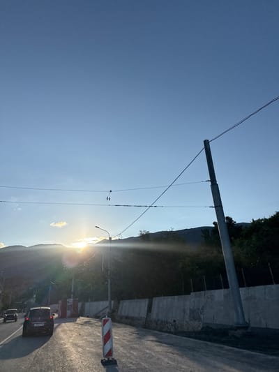 The image depicts a roadside scene at sunset, with mountains in the background and a clear blue sky overhead. The sun is low on the horizon, casting a warm glow and creating lens flares. A road runs diagonally across the lower portion of the frame, with two cars visible: a black SUV in the foreground and a darker vehicle further back. The road appears to be under construction, with a concrete barrier and a red and white striped traffic cone visible on the right side. A tall utility pole with wires stretches across the frame. The setting appears to be a rural or suburban area, possibly on the outskirts of Pushkino, Ukraine, given the natural landscape and the presence of basic infrastructure.
