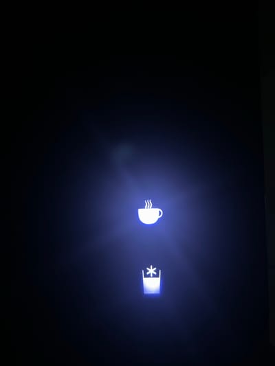 The image depicts two illuminated symbols on a dark background. On the left is a white outline of a coffee cup with emanating steam lines. To the right of the cup, there's a rectangular symbol with a white asterisk inside it. There is no evidence to connect the image to Cheongju-si, South Korea. No people, objects, actions, interactions, or environmental details beyond the symbols and background can be determined from the image.