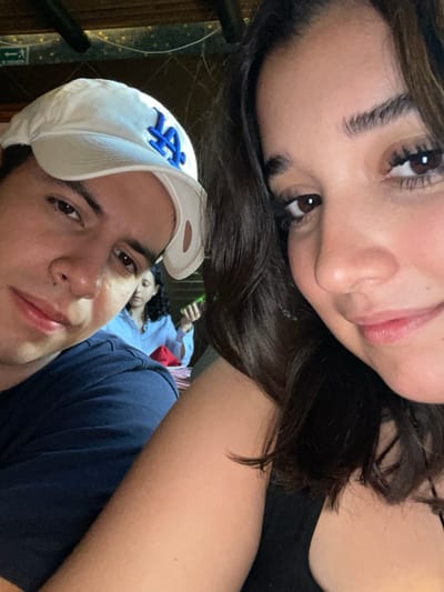 The image presents a selfie-style portrait of three individuals within an indoor restaurant setting in Boca del Río, Mexico.
In the foreground, a female with dark, wavy hair, dark eyes, and fair skin occupies the right two-thirds of the frame. She wears a dark-colored top and looks towards the camera with a subtle smile. To her left, partially obscured and positioned slightly behind, is a male with dark hair, dark eyes, and fair skin. He wears a white baseball cap with a blue "LA" logo and a dark blue t-shirt, looking directly at the camera with a neutral expression.
In the middle ground, behind the two foreground subjects, a third female with dark, curly hair is seated at a table. She wears a light-colored, possibly striped, collared shirt. Her right hand holds a green, elongated object. A red napkin is visible on the table in front of her.
The background consists of dark wooden structural beams and a lighter-colored wall or ceiling section. A red "EXIT" sign with a white arrow is partially visible in the upper left portion of the background.