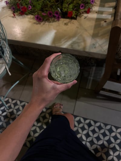 A person's hand holds a clear glass filled with a light-colored beverage and ice cubes. The glass is transparent and has a faceted design. The hand is fair-skinned with visible veins and is positioned in the lower right quadrant of the image, close to the camera. The person is wearing a dark blue garment, possibly a dress or skirt, and a sandal on their right foot is visible in the lower right corner. The background features a tiled floor with a black and white geometric pattern. To the left, a portion of a light blue metal chair is visible. In the upper part of the image, a planter with green foliage and red and purple flowers can be seen. The scene appears to be outdoors, possibly on a patio or in a garden setting, with artificial lighting casting shadows. The location is Beit Hanan, Israel.
