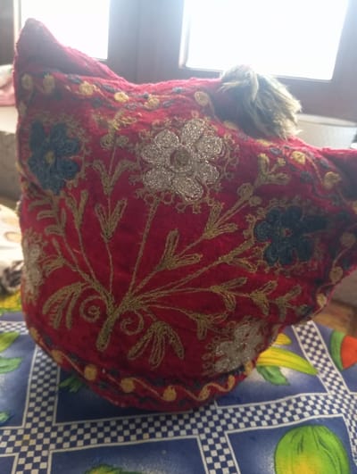 A close-up, slightly blurry, eye-level shot shows a red, intricately embroidered hat on a blue and white checkered tablecloth. The hat is adorned with gold and blue floral patterns and has a tuft of light brown fur sticking out from the top. The background is out of focus, with a window letting in bright light. The scene appears to be indoors, on a table with a patterned tablecloth. No specific location in Chumbagish, Uzbekistan is identifiable.