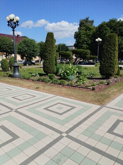 The image depicts a bright, sunny day in a park or public square, likely in Barysaw, Belarus. The foreground is dominated by a decorative paved walkway with a geometric pattern of light gray and pale green square tiles. Beyond the walkway, a landscaped garden area features various shrubs and plants, including a large cluster of white-flowering hydrangeas. Several tall, conical evergreen trees and rounded, manicured bushes are strategically placed within the garden. A lamppost with five globe-shaped lights stands to the left, while another similar lamppost is visible in the mid-ground to the right. In the background, a building with a reddish-brown roof and light-colored walls is partially visible, along with more trees and a glimpse of parked cars and a few people sitting on a bench. The sky is a vibrant blue with scattered white, puffy clouds, suggesting pleasant weather. The overall impression is one of a well-maintained, public outdoor space on a clear summer day. There is no visible text in the image.