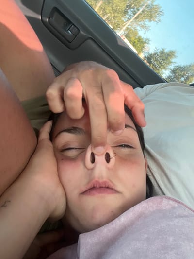 A fair-skinned person with dark hair and a nose ring reclines, eyes closed, head resting on their hand. Another person's hand pinches their nose. They appear to be in a car, with a window and door panel visible on the left. The person is lying against a white pillow with a lavender-colored blanket nearby. A small tattoo is on the wrist of the hand supporting the head.
