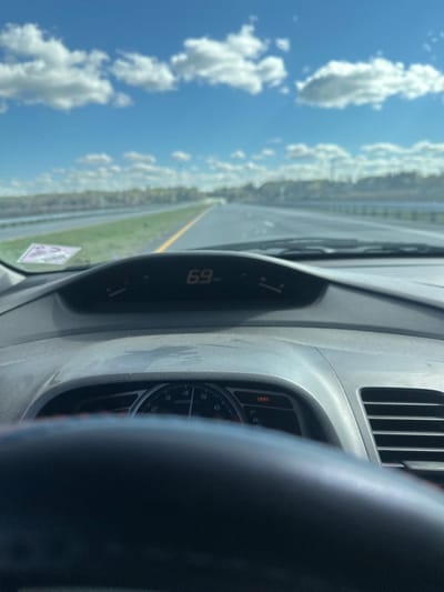 This image, captured from the driver's perspective inside a vehicle, depicts a multi-lane highway under clear blue skies with scattered white cumulus clouds. The vehicle is in motion, with the digital speedometer on the upper dashboard displaying "69", indicating a speed of 69 miles per hour. The lower dashboard shows a tachometer with a needle resting between 1000 and 2000 RPM, and an illuminated red warning light on the right, consistent with a Tire Pressure Monitoring System (TPMS) alert. The dashboard surfaces appear dusty. A purple-hued sticker, possibly a permit or toll tag, is affixed to the lower left portion of the windshield.
The external environment visible through the windshield shows a paved asphalt highway with a solid yellow line delineating the left edge of the travel lane. Green grassy areas and distant trees line both sides of the highway, with what may be a body of water or reflective surface visible beyond the median on the left. The scene is consistent with travel on a major highway, such as Interstate 75, in or near Gainesville, Florida, United States, during daylight hours. No individuals are visible in the frame.