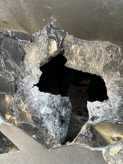 The image displays a close-up, top-down view of a large, irregular hole in a shattered, translucent panel, consistent with broken tempered glass. The fragmentation pattern exhibits numerous small, interconnected, crystalline-like shards with sharp, jagged edges, heavily refracting and reflecting light. The central aperture, roughly amoeboid in shape, reveals a dark, indistinct void or interior space beyond. Portions of a lighter, opaque structural frame or mounting surface are visible at the periphery of the shattered material, particularly at the top and bottom of the frame. No individuals, discernible objects, or specific actions are present within the immediate scene. This damage is located in the city of Dura, Palestinian Territory.