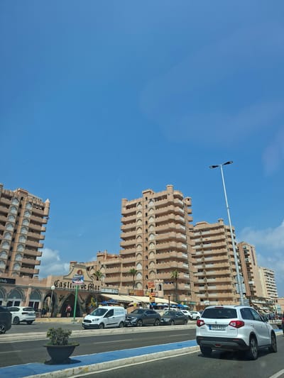 The image depicts a street scene in front of a building complex named "Castillo de Mar" in San Javier, Spain. The buildings are tall, multi-story structures with a distinctive architectural style featuring arched windows and crenellated parapets. They are primarily a light brown or beige color. In the foreground, a road with blue and white markings is visible, with several cars driving on it, including a white Suzuki Vitara with license plate "3940 KDG". There is a potted plant on the left side of the road. The street level features various businesses and shops with awnings and signs, including one that says "MaleniJet" and another that appears to be "Marrakech Bazaar". The sky above is clear and blue with some scattered white clouds. The overall scene suggests a commercial and residential area by the coast.
