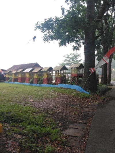 The image displays an outdoor scene in Cugenang, Indonesia, under an overcast sky. In the mid-ground, a linear arrangement of approximately eight elevated wooden huts or gazebos with dark, peaked roofs and red and yellow painted base structures extends horizontally. Behind these, to the left, a larger building with a multi-peaked, dark-tiled roof is partially visible. A low, blue-painted retaining wall or curb runs along the front of the huts. The foreground consists of damp green grass, interspersed with patches of brown earth and scattered dead leaves. To the right, an asphalt or paved path curves slightly upwards, bordered by several irregularly shaped concrete or stone slabs embedded in the ground. Multiple large trees with dark trunks and dense green canopies occupy the right side of the frame. A red and white striped flag is wrapped around the trunk of the most prominent tree on the right. In the distant background, beneath the bright, overcast sky, a lighter green coniferous tree stands out. A small, dark, airborne object with a tail is visible against the sky in the upper left quadrant. No individuals or animals are present in the scene. The overall environment appears damp, indicating recent precipitation.