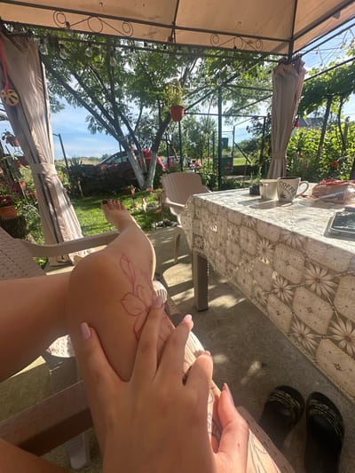 The image depicts a sunlit outdoor patio with a woman's legs and arms in the foreground. She is seated, with one leg extended and a temporary tattoo of a flower on her thigh. Her hand is visible, with neatly manicured fingernails. In the background, a table is set with two coffee mugs, one of which says "IT'S COFFEE TIME," and a package of cookies. The patio is covered by a canopy, and plants are visible in pots and growing around the area. The background shows a garden with trees, a fence, and a red car. The scene appears to be a relaxed outdoor setting. The location is identified as Rogozen, Bulgaria.
