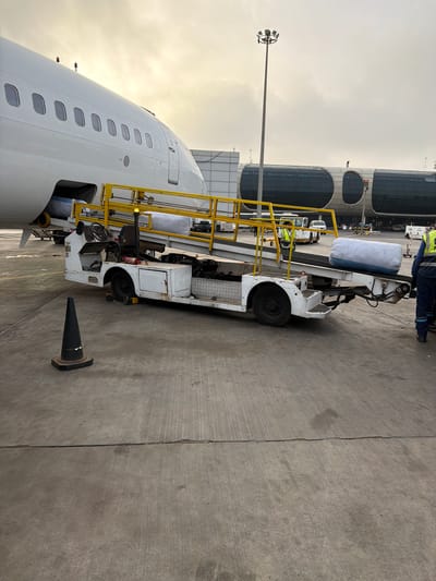 The image shows the exterior of an airport in Communauté rurale de Diass, Senegal. A white passenger airplane is parked on the tarmac. A white luggage carrier with yellow railings is parked alongside the airplane. A black traffic cone stands nearby on the concrete. An airport terminal is visible in the background. The sky is overcast. An airport light pole is visible in the background. A person wearing a neon yellow vest is partially visible on the luggage carrier.
