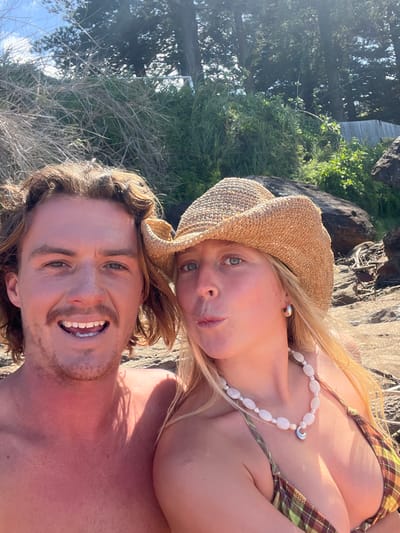 A close-up selfie captured at an unspecified coastal location in Coalcliff, Australia, shows two individuals. On the left, a male with shoulder-length brown hair and a light brown mustache and goatee smiles, displaying upper and lower teeth. His chest and neck are bare and appear sunburned. His left shoulder is visible in the lower left foreground. On the right, a female with blonde hair wears a straw cowboy hat. Her blue eyes are visible, and her lips are pursed as if blowing a kiss. She wears a multi-strand necklace of white, irregular, oval-shaped beads interspersed with smaller round pearls, featuring a crescent moon pendant. Her attire includes a plaid bikini top in shades of green, brown, and yellow. A silver hoop earring is visible in her right ear.
The background reveals a sunlit, rugged coastal environment. Behind the subjects, dark brown rocks and patches of green and dry brown vegetation are visible. Further back, tall, dark green trees stand against a clear, light blue sky with sparse white clouds. A glimpse of a light-colored fence or structure can be seen through the foliage in the upper background. Sunlight casts distinct shadows on the male's neck and chest, indicating direct overhead lighting.