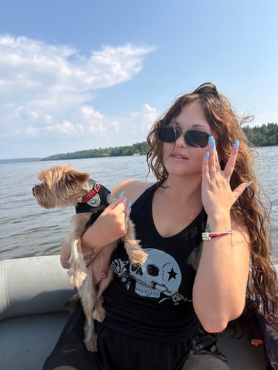 This image, taken in the city of Sylva, Russia, depicts a young woman holding a small dog while on a boat. The woman, with long, wavy brown hair, is wearing a black tank top with a skull graphic, dark sunglasses, and has long, light blue painted fingernails. She is touching her face with her right hand. The dog, a Yorkshire Terrier, is wearing a black harness with a red collar. The boat appears to be an inflatable raft, visible on the left side of the frame. The background shows a body of water, a tree-lined shore, and a blue sky with scattered clouds.