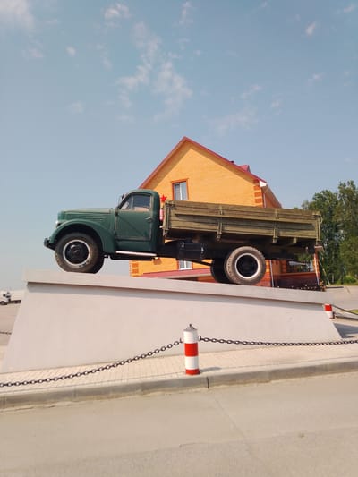 In Krasny Yar, Russia, an old, dark green truck is displayed prominently on a white, elevated platform. The truck is positioned facing left, with its front wheel elevated and its rear wheels on the platform. The truck features a wooden cargo bed and a red star emblem on its side. Behind the truck, a building with orange brick walls and a red roof is visible. The sky above is a clear blue with scattered white clouds. A chain barrier with red and white striped posts separates the platform from a paved area in front of it. The scene appears to be an outdoor display or monument.