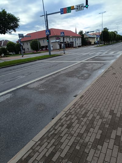 The image captures a view of a street in Jõhvi, Estonia, likely near a prominent intersection. A traffic light showing a green signal is visible, indicating clear passage. Pedestrian crossing signs are present, with a white silhouette of a person walking on a blue background. Buildings with red roofs line the street, featuring architectural details suggestive of public or commercial establishments. The road surface appears wet, possibly from recent rain, with reflections visible on the asphalt. A brick-patterned sidewalk runs along the right side of the road. Trees are planted along the street, some with full foliage, contributing to the urban landscape. The sky is overcast with clouds. The overall scene depicts a typical day in a moderately urbanized area.