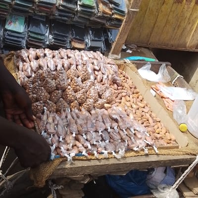 An elevated view captures a market stall in Olomoda, Nigeria, displaying various packaged goods. The primary subject is an inclined wooden surface, covered with burlap, laden with brown, nut-like items. A large quantity of these items are loose, forming a heap, while others are individually packaged in small, clear plastic bags tied with white string, arranged in orderly rows. A central pile consists of larger, irregularly shaped items, also clear-bagged. Two dark-skinned hands are visible on the left, positioned as if arranging the merchandise on the stall.
To the right of the main display, a small pile of white granular substance is contained within a clear plastic bag, alongside a larger white plastic bag and an empty clear plastic water bottle with a yellow cap. Above these items, a bright blue object rests on a wooden support.
The background features a vertical display wall, lined with numerous small, flat, rectangular items, individually sealed in clear plastic, resembling phone cases or wallets, stacked in rows. The stall structure consists of rough wooden planks. Beneath the main display, a blue plastic bag and other indeterminate objects are visible, resting on an uneven surface.