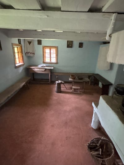 This image depicts the interior of a rustic room, likely a dwelling in Sielachowskie, Poland, given the context. The scene appears to be a historical or traditional setting, possibly a museum exhibit or a preserved old home. The room is sparsely furnished, with an emphasis on functional, rustic objects.

Dominating the space are wooden beams supporting the ceiling and framing the structure of the room. The walls are a light blue, and the floor is a reddish-brown earthen material, suggesting a simple and traditional construction.

Several windows with wooden frames and panes are visible. One larger window on the left shows a grid pattern of smaller panes, letting in natural light. Another smaller window is visible on the right.

Objects in the room include a dark wooden table or work surface with various items on it, including what appears to be a barrel and some metal implements. A basket filled with sticks or kindling sits on the floor near a white object that might be a bag or cloth. There are also framed pictures on the wall, adding a touch of personal history to the space. A piece of embroidered fabric, possibly a wall hanging or tablecloth, with red floral patterns and a triangular motif, is draped over something on the wall.

The lighting suggests it is daytime, with light streaming in through the windows. The overall atmosphere is one of simplicity, history, and perhaps a sense of quietude, evoking a past way of life. There is no visible text in the image.