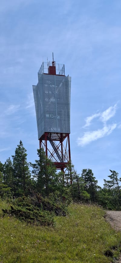 A tall, modern red and white tower stands against a clear blue sky with scattered white clouds. The tower has a red metal frame supporting a white, slatted structure that flares outwards at the top. A set of metal stairs leads up the side of the tower. The tower is situated on a rocky, grassy hilltop dotted with pine trees and dense green bushes. In the foreground, a field of green grass with small wildflowers is visible. This scene appears to be of the Panga cliff on the island of Saaremaa, near Panga village, Estonia, a popular tourist spot known for its scenic views. There are no people visible in the image.