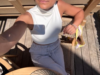 A woman is seated in a woven chair on a wooden deck, possibly in Ramatuelle, France. She is wearing striped pants and a white top. A hat sits on the table nearby. The scene is brightly lit, suggesting it is daytime.