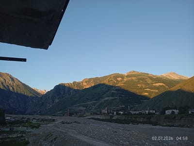 The image depicts a scenic mountain valley, likely in or near the city of Tyrnyauz, Russia, bathed in the golden light of early morning. The foreground is a rocky, dry riverbed or valley floor with a dirt road winding through it. Sparse vegetation and small structures, possibly remnants of construction or dwellings, are scattered on the left side of the valley floor.
Dominating the mid-ground and background are majestic, verdant mountains. The slopes of these mountains are terraced in some areas, suggesting human activity or natural formations. Shadows cast by the mountains stretch across the landscape, indicating the low angle of the sun.
A small settlement is visible nestled at the base of the mountains, featuring several buildings, some of which appear to be residential or industrial. Two prominent radio towers, one red and white and another red, stand out against the mountain backdrop. Power lines and poles are also visible, traversing the mountain slopes.
In the far distance, snow-capped peaks of higher mountains are visible, their summits catching the sunlight. The sky is a clear, bright blue, devoid of clouds.
In the extreme foreground, a dark, angular concrete structure, possibly part of a bridge or overpass, partially obscures the view. A thin metal beam extends horizontally from this structure.
The timestamp "02.07.2026 04:58" is overlaid on the lower right portion of the image, indicating the date and time the photograph was taken. No people are discernible in the image. The overall impression is one of a remote, mountainous landscape at dawn, with signs of human habitation and infrastructure integrated into the natural environment.