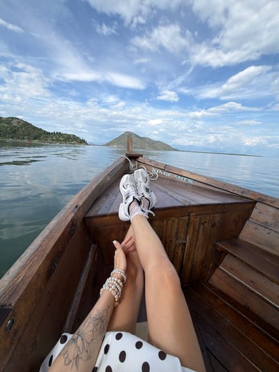 A person is sitting on a wooden boat, with their legs crossed. They are wearing white sneakers and a dress with black polka dots. The person has pearl bracelets on their left wrist, and a tattoo of a flower on their left forearm. The boat is on a body of water, possibly a lake, with a mountain range in the background. The sky is blue with white clouds. The words "KINGF SHFER" are visible on the side of the boat. The location is in the city of Braćeni, Montenegro.